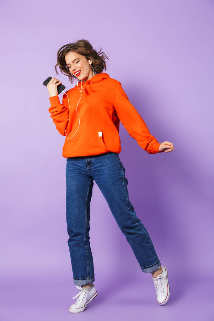 Image of young woman isolated over purple background wall listening music with earphones holding phone. - Photo, Image