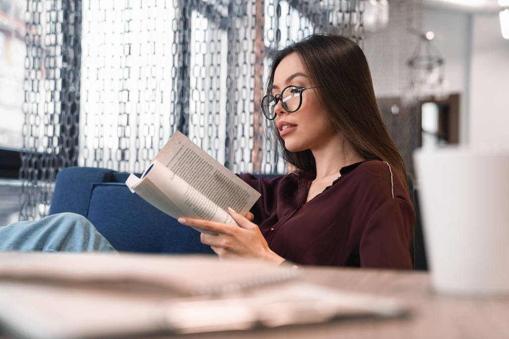 Aantrekkelijke vrouw in brillen lezen boek bij de caf cup en documenten op voorgrond - Foto, afbeelding