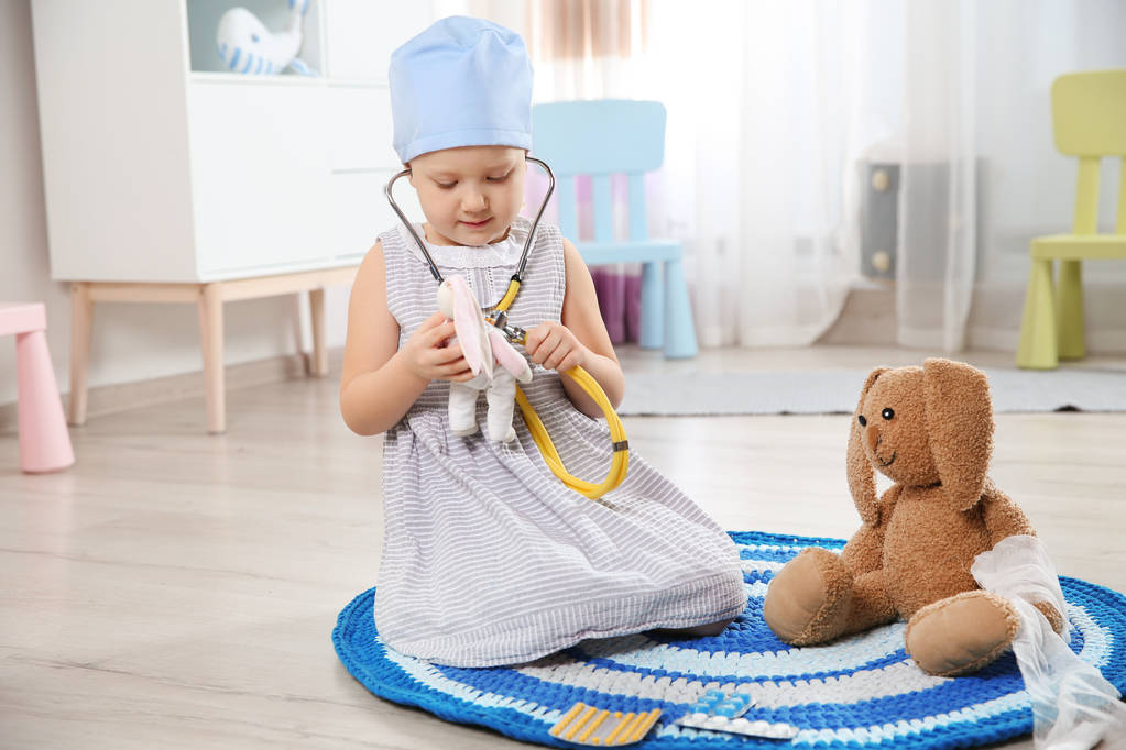Söpö lapsi kuvittelee itsensä lääkäriksi leikkiessään stetoskoopilla ja lelupupulla kotona
 - Valokuva, kuva