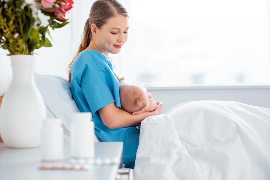 vista lateral de la feliz madre joven sentada en la cama y amamantando al bebé recién nacido en la habitación del hospital
 - Foto, imagen