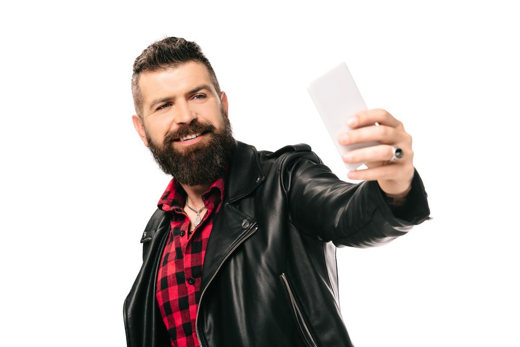Lächelnder bärtiger Mann in schwarzer Lederjacke macht Selfie auf Smartphone, isoliert auf weißem Hintergrund - Foto, Bild