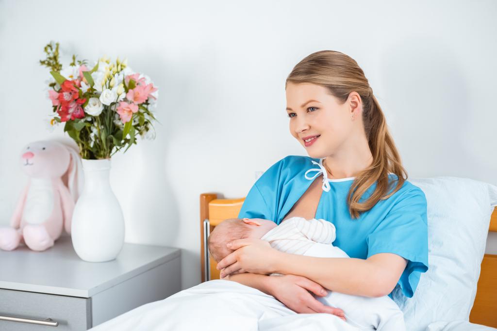 happy young nursing mother breastfeeding newborn baby and looking away in hospital room - Photo, Image