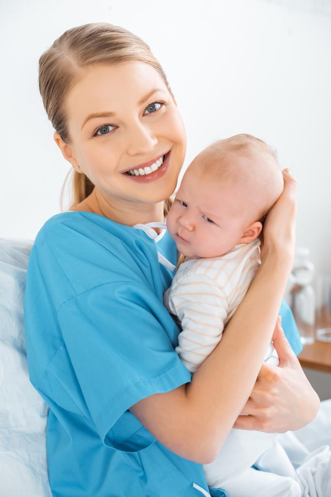 feliz jovem mãe sentada na cama com bebê adorável e sorrindo para a câmera no quarto do hospital
 - Foto, Imagem