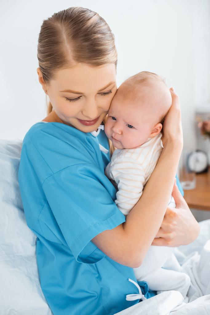 glückliche junge Mutter sitzt auf Krankenhausbett und umarmt entzückendes Baby - Foto, Bild