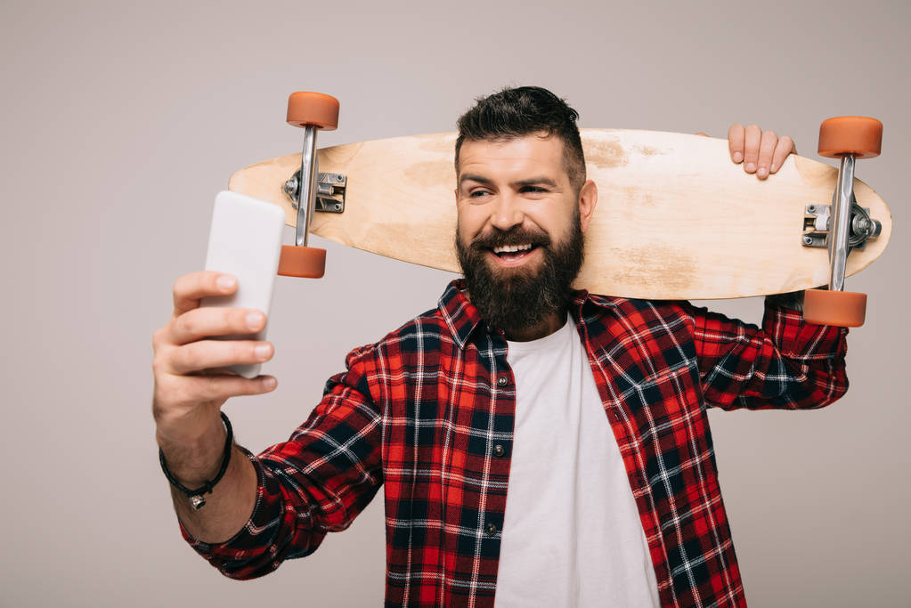 cheerful man in checkered shirt holding longboard and taking selfie on smartphone isolated on grey - Photo, Image