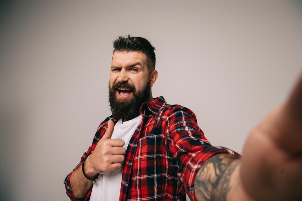 excited man in checkered shirt showing thumb up while taking selfie, isolated on grey - Photo, Image