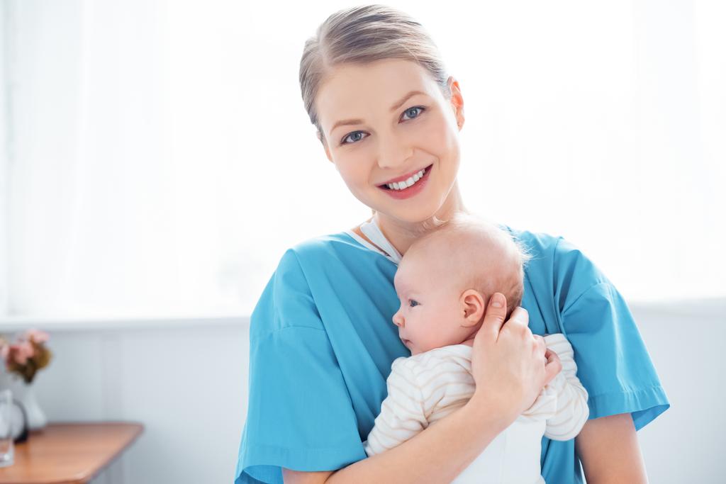 onnellinen nuori äiti tilalla vastasyntynyt vauva ja hymyilee kameran sairaalahuoneessa
 - Valokuva, kuva