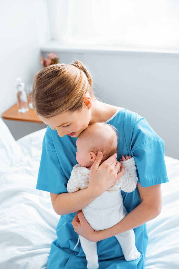 vista de ángulo alto de feliz madre joven sentada en la cama y sosteniendo al bebé recién nacido en la habitación del hospital
  - Foto, imagen
