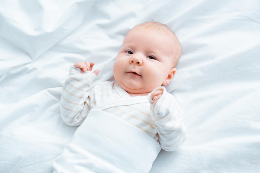 top view of adorable baby looking at camera while lying on white bedding  - Photo, Image