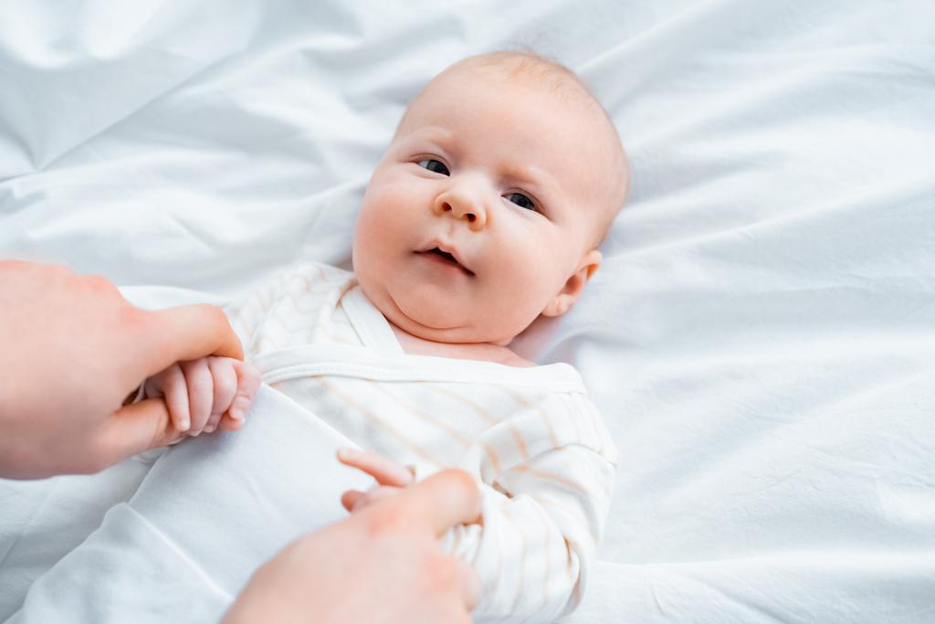 tiro recortado de la madre cogida de la mano del bebé adorable acostado en la ropa de cama blanca
 - Foto, Imagen