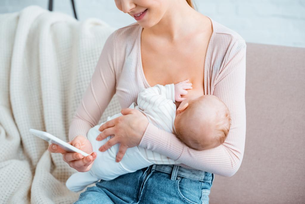 περικοπεί shot χαμογελώντας νεαρή μητέρα θηλάζει μωρό και χρησιμοποιώντας το smartphone στο σπίτι - Φωτογραφία, εικόνα