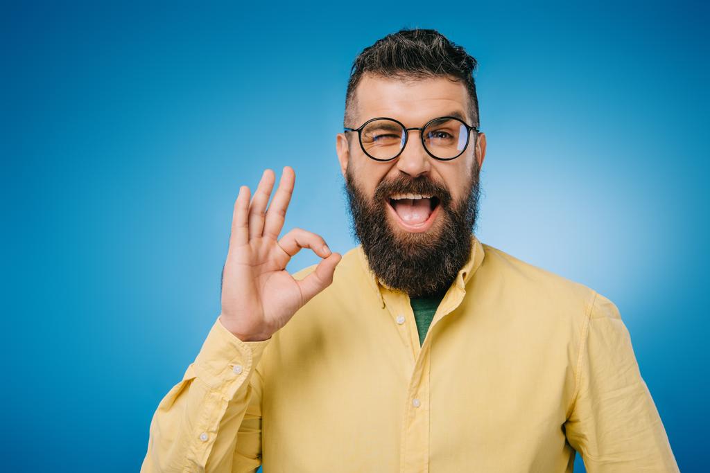 счастливый бородатый мужчина в очках подмигивает и показывает знак "ОК", изолированный на голубом фоне
 - Фото, изображение