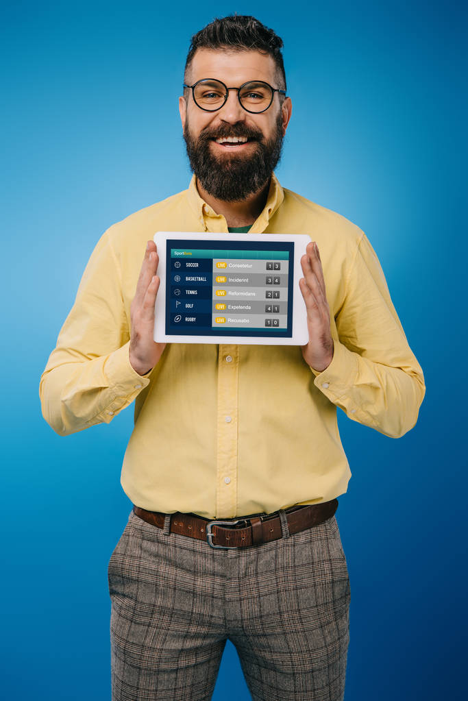 Lächelnder bärtiger Mann zeigt digitales Tablet mit Sport-App, isoliert auf blauem Grund - Foto, Bild