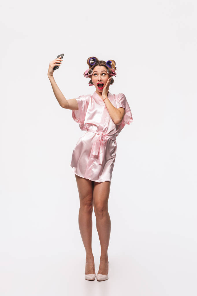 Volledige lengte portret van een mooie huisvrouw met krulspelden in haar haren kleed staande geïsoleerd op een witte achtergrond, dragen het nemen van een selfie met mobiele telefoon - Foto, afbeelding