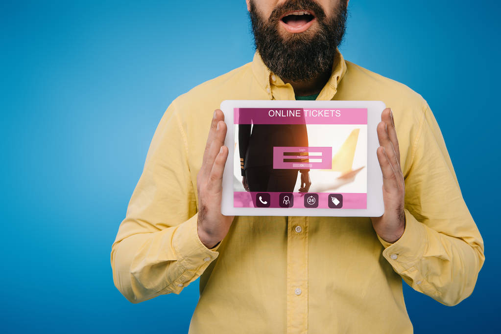 Ausgeschnittene Ansicht eines schockierten bärtigen Mannes, der ein digitales Tablet mit Online-Ticket-App zeigt, isoliert auf blauem Grund - Foto, Bild