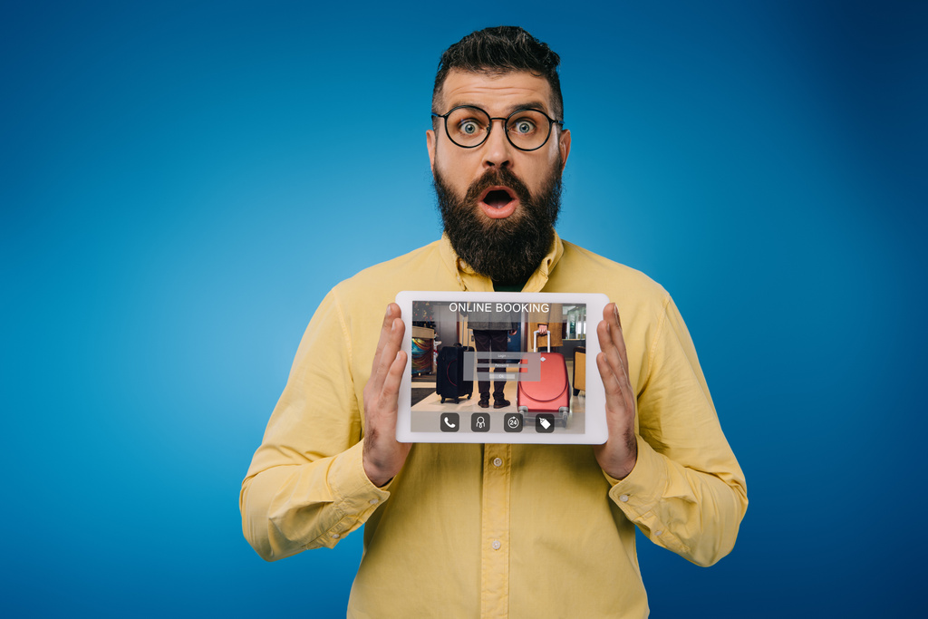 Šokovaný vousatý muž představují digitální tabletu s on-line rezervace app, izolované na modré - Fotografie, Obrázek