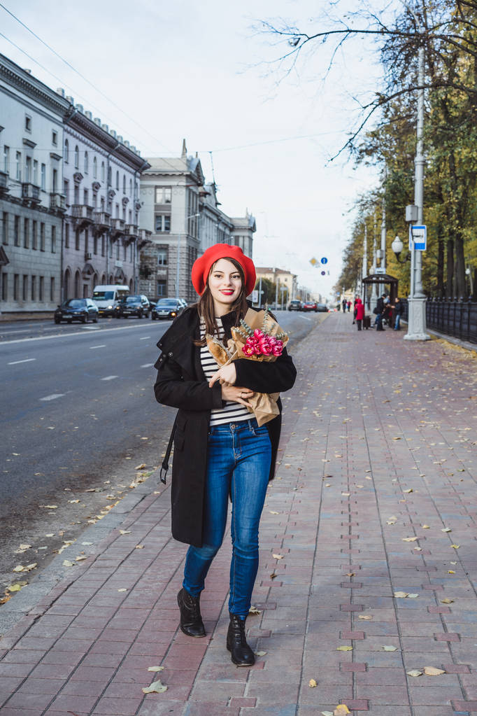 Красива дівчина брюнетка Оттона в червоний бере і чорні пальто йде по вулиці європейського міста з букетом квітів і французький багети. Щасливою усмішкою і чарівною дивитися, французькому стилі Парижа - Фото, зображення