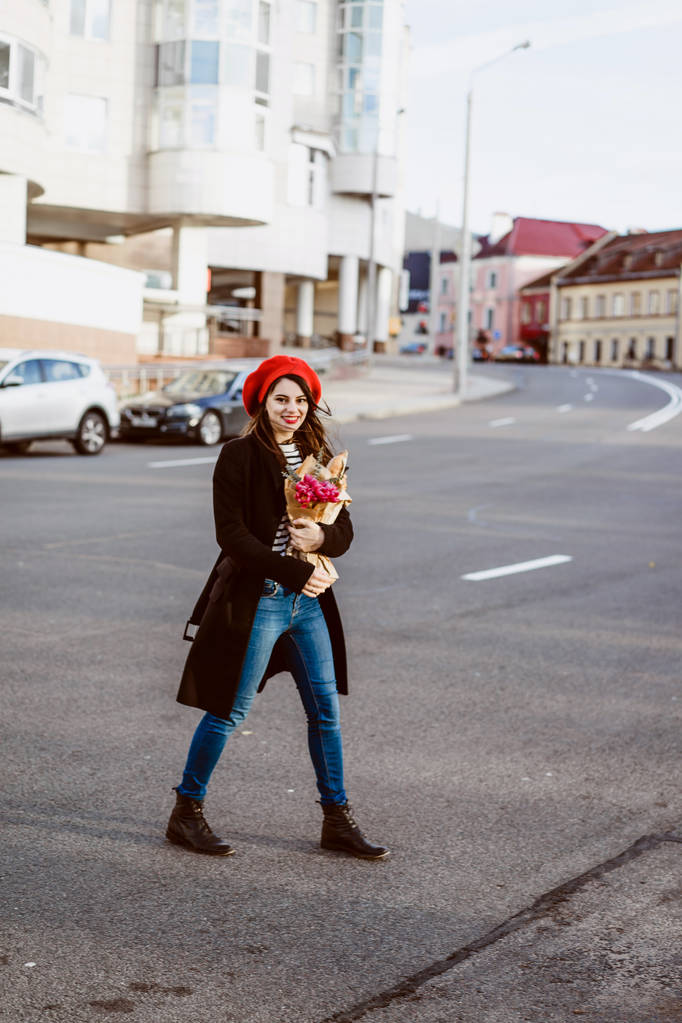 Mooi meisje Française brunette in een rode baret en een zwarte jas gaat langs de straat van de Europese stad met een boeket van bloemen en Franse stokbrood. Een tevreden glimlach en een charmante look, de Franse stijl van Parijs - Foto, afbeelding