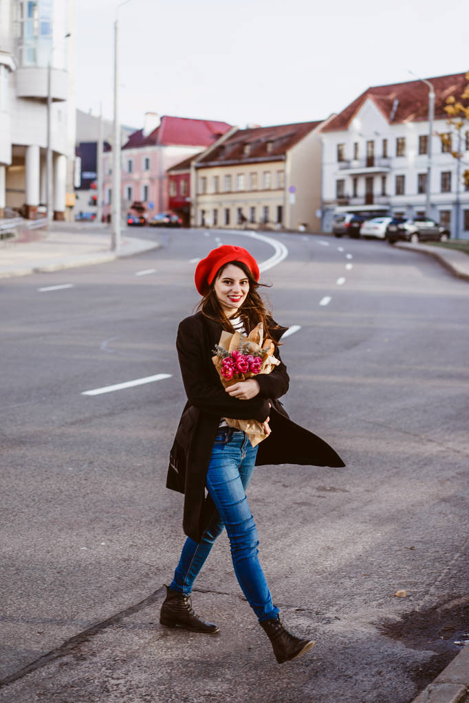 Fiatal gyönyörű lány francia nő barna-piros beret és egy fekete kabát megy az utcán, az európai város, egy csokor virágot és francia bagettet. A boldog mosoly és egy bájos megjelenés, a francia stílusú, Párizs - Fotó, kép