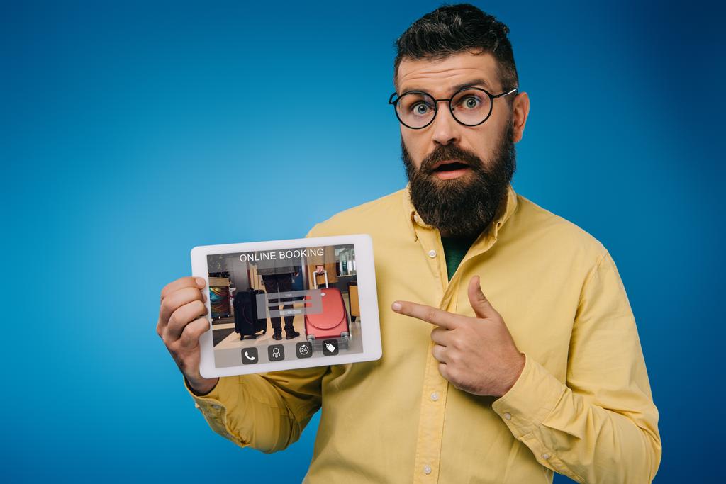 шокований бородатий чоловік вказує на цифровий планшет з додатком онлайн-бронювання, ізольований на синьому
 - Фото, зображення