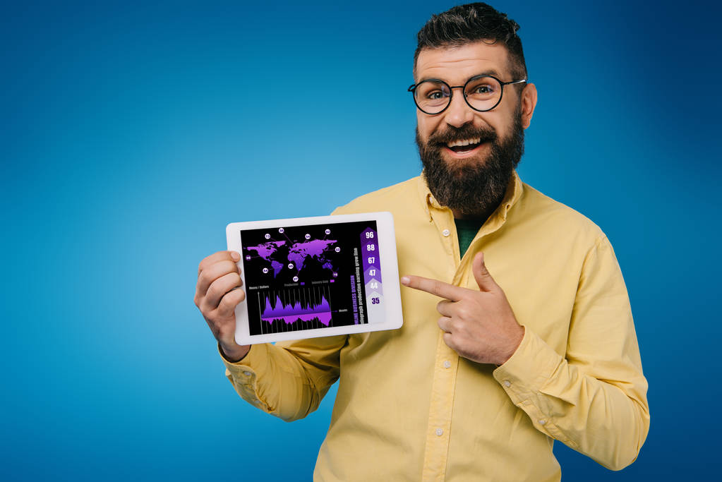 Lächelnder bärtiger Mann zeigt auf digitales Tablet mit Infografik, isoliert auf blauem Grund - Foto, Bild