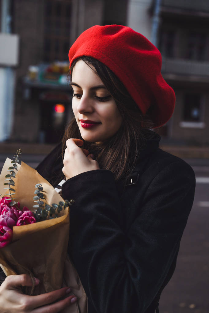 Krásná mladá dívka Francouzka brunetka v červený baret a Černý kabát jde po ulici Evropská města s kyticí květin a francouzské bagety. Šťastný úsměv a okouzlující vzhled, francouzském stylu Paříže - Fotografie, Obrázek