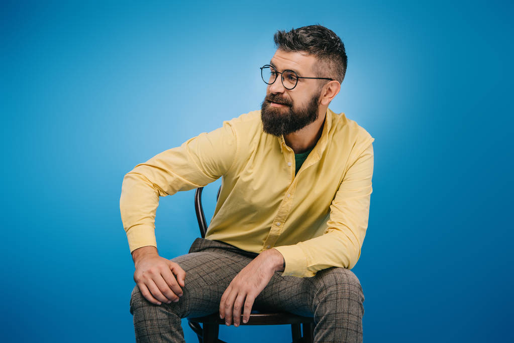 pohledný muž v brýlích, sedí na židli, samostatný na modré - Fotografie, Obrázek