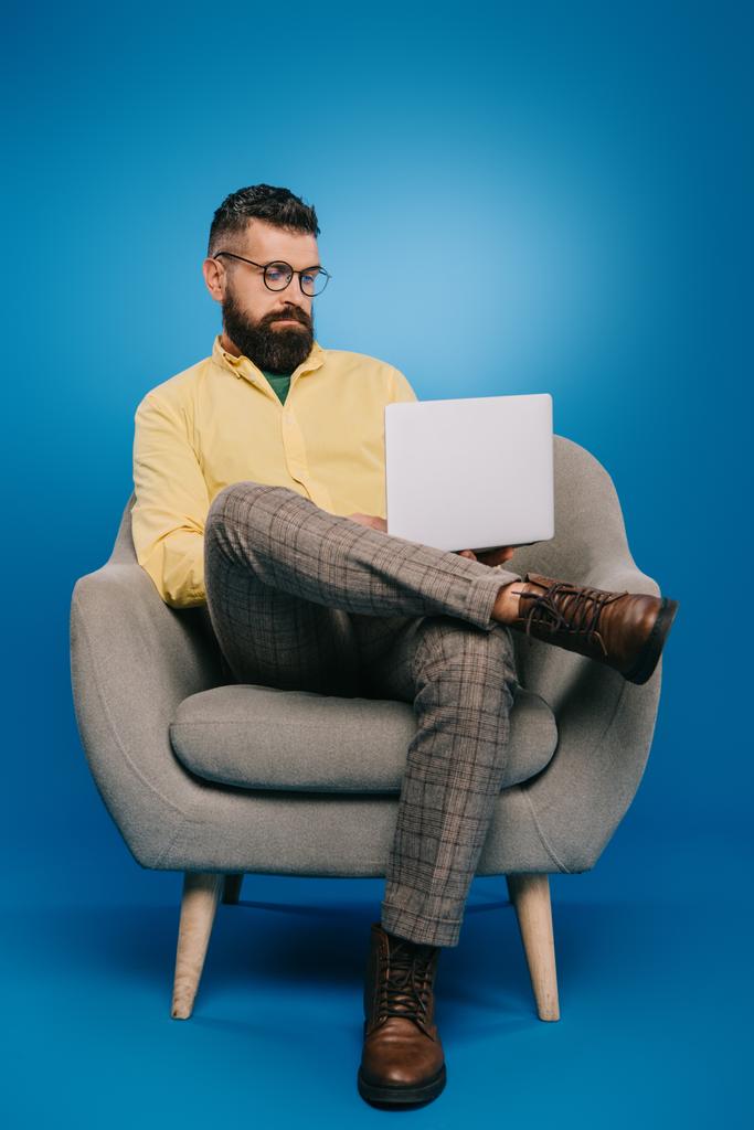 όμορφος γενειοφόρος επιχειρηματία που κάθεται στην πολυθρόνα με το laptop στο μπλε - Φωτογραφία, εικόνα