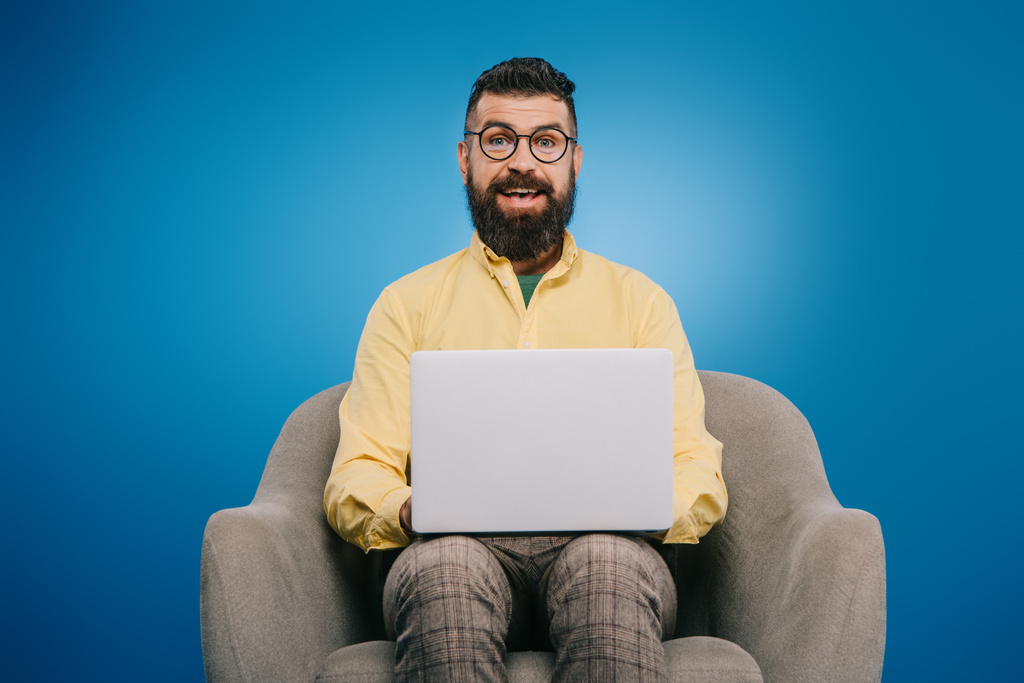 ευτυχισμένος γενειοφόρος επιχειρηματίας χρησιμοποιώντας φορητό υπολογιστή ενώ κάθεται στην πολυθρόνα, απομονώνονται σε μπλε  - Φωτογραφία, εικόνα