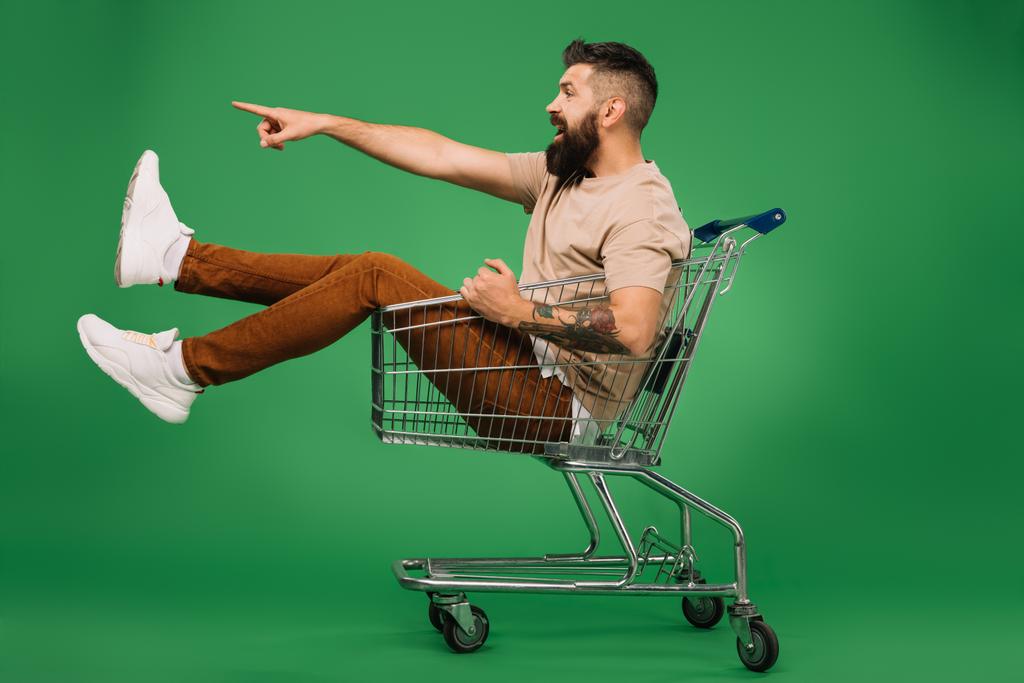 збуджений бородатий чоловік показує щось, сидячи в кошику для покупок ізольовано на зеленому
 - Фото, зображення