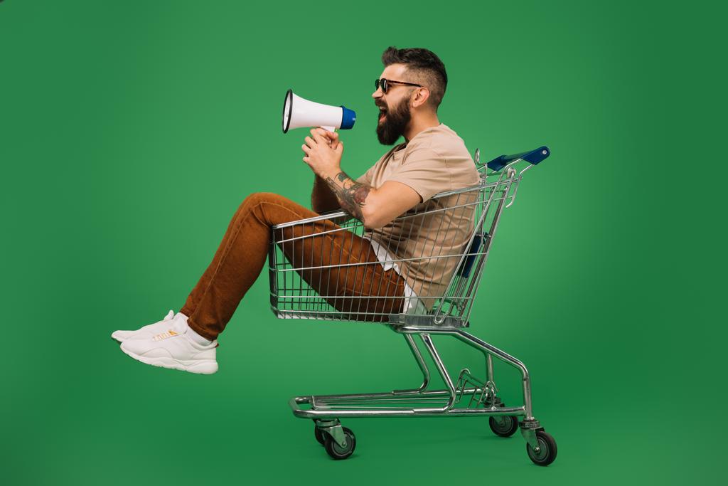 barbudo gritando en megáfono mientras está sentado en el carro de la compra aislado en verde
 - Foto, imagen