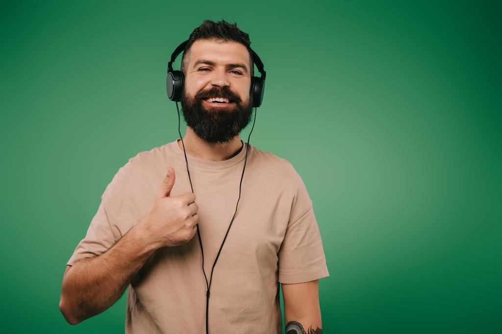 bearded smiling man showing thumb up while listening music in headphones isolated on green - Photo, Image