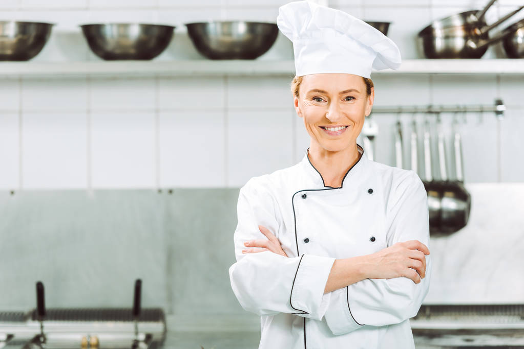 kaunis hymyilevä naiskokki univormussa ja hattu lwith kädet ristissä ooking kameran ravintolan keittiössä
 - Valokuva, kuva