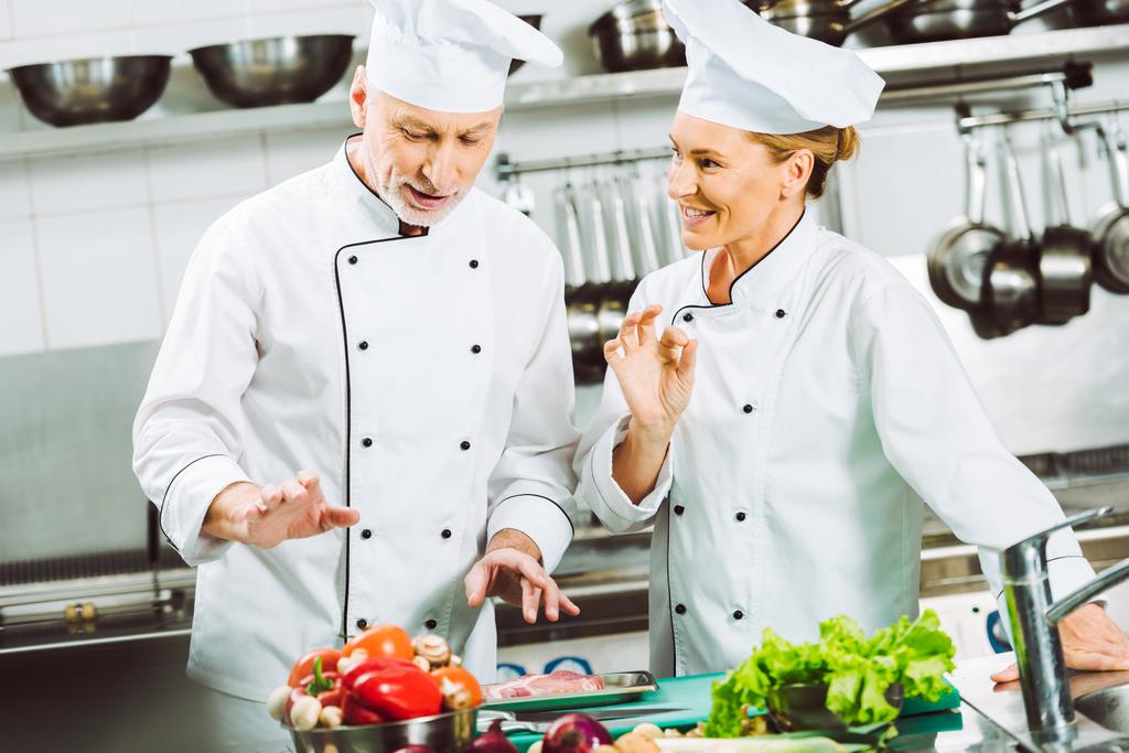 άνδρες και γυναίκες σεφ στην ομοιόμορφη και καπέλα να μιλάμε, ενώ το μαγείρεμα στην κουζίνα εστιατορίου - Φωτογραφία, εικόνα