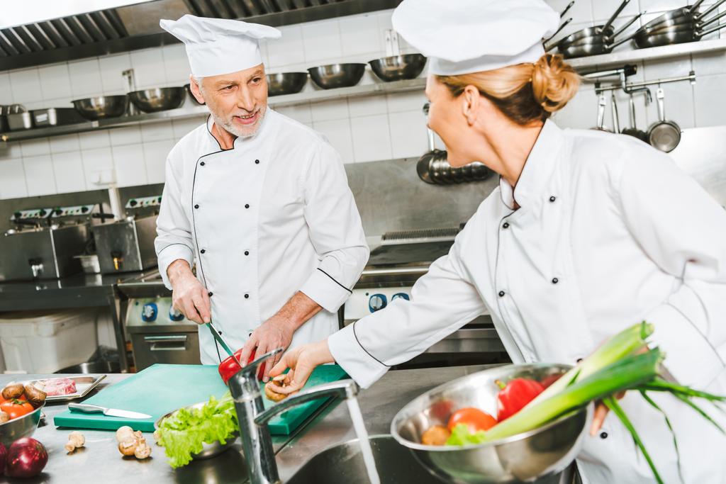 mannelijke en vrouwelijke chef-koks in uniform en hoeden kijken elkaar tijdens het koken in de keuken van het restaurant - Foto, afbeelding