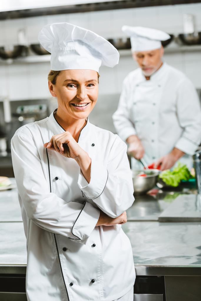 kaunis hymyilevä naiskokki univormussa ja hattu katselee kameraa ravintolan keittiössä
 - Valokuva, kuva