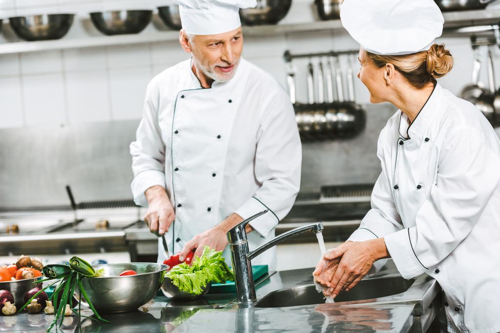 selective focus of female and male chefs in uniform looking at each other while cooking in restaurant kitchen - Photo, Image