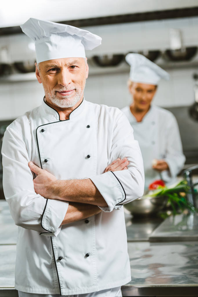 schöner männlicher Koch in Uniform mit verschränkten Armen, der in der Restaurantküche in die Kamera blickt - Foto, Bild