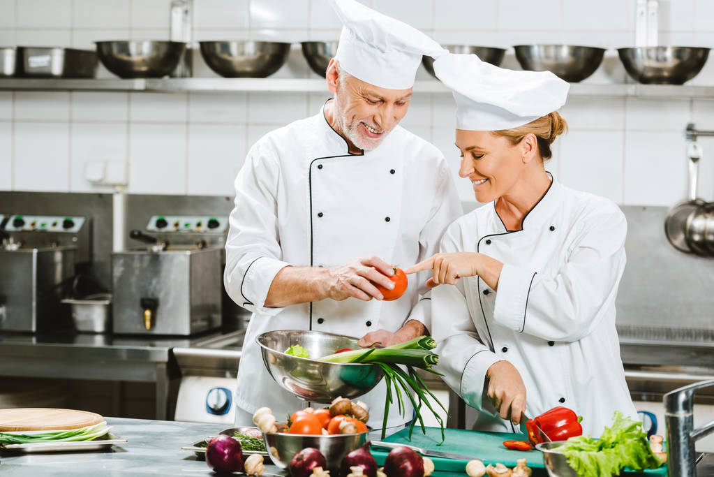 lachende mannelijke en vrouwelijke chef-koks in double-breasted jassen en hoeden koken in de keuken van het restaurant - Foto, afbeelding