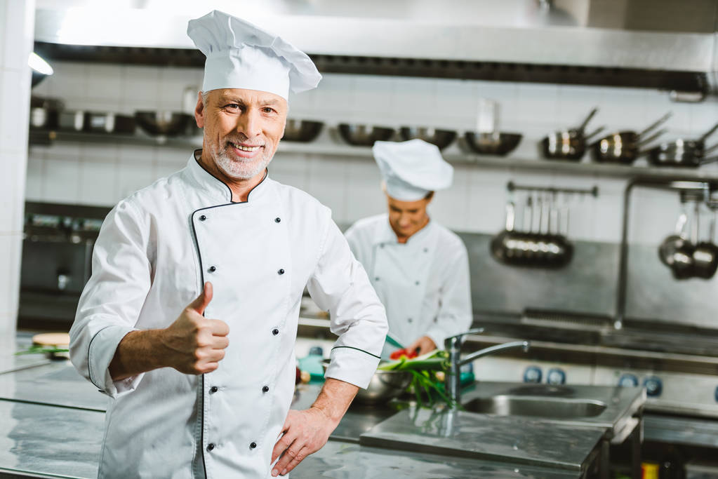 hezký, usmívající se muž kuchař v uniformě při pohledu na fotoaparát a ukazuje palcem podepsat v kuchyni restaurace - Fotografie, Obrázek