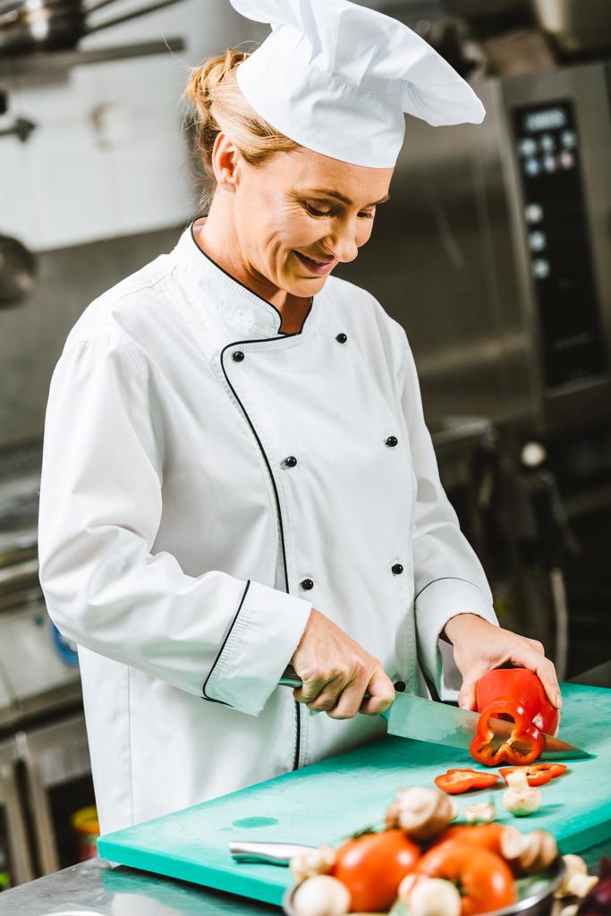 krásné, usmívající se žena kuchař v jednotných stříhání pepř v kuchyni restaurace - Fotografie, Obrázek