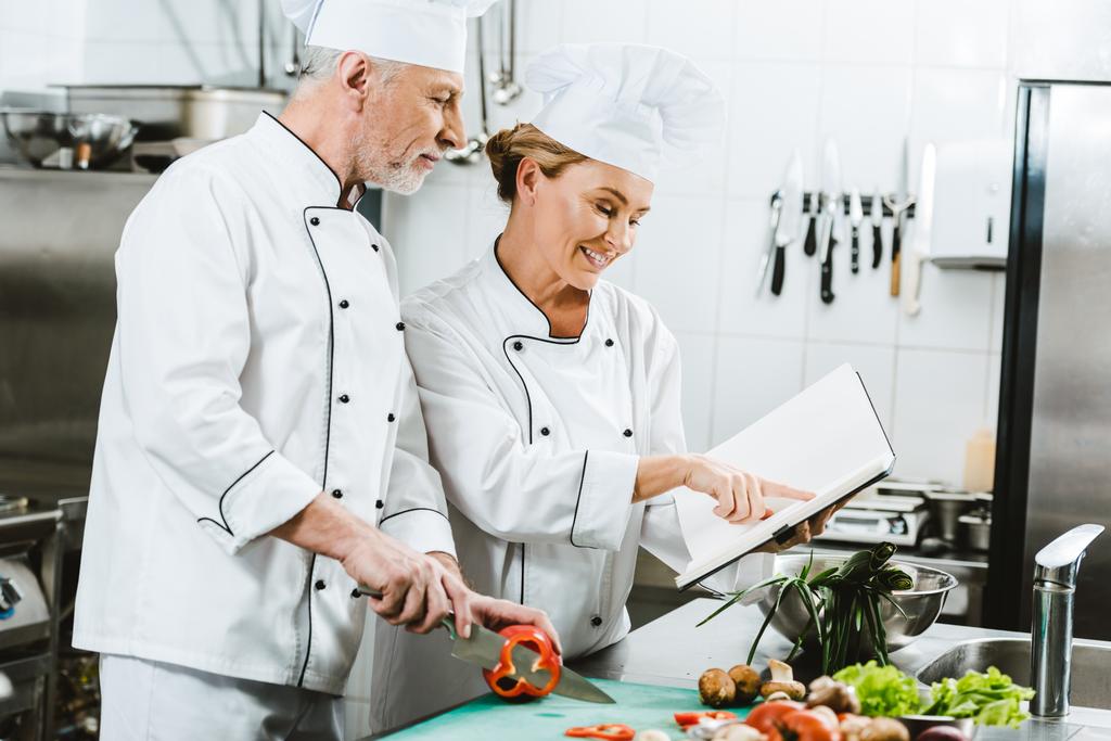 selective focus of female and male chefs in uniform using recipe book during cooking in restaurant kitchen - Photo, Image