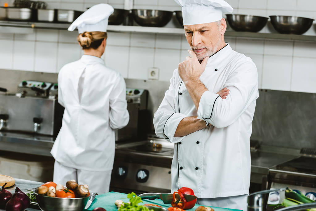 背景の女性の同僚とレストランの厨房で調理中にダブル ブレストのジャケットで物思いにふける男性シェフ - 写真・画像