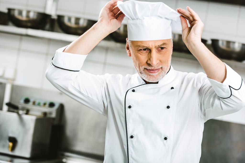 bonito masculino chef no uniforme ajustando cap no restaurante cozinha
 - Foto, Imagem
