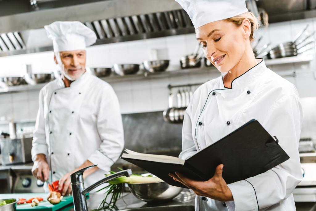 vrouwelijke chef-kok in uniform lezen receptenboek terwijl collega koken op achtergrond in restaurant keuken - Foto, afbeelding