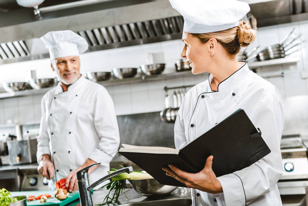 женщина шеф-повар в униформе держит книгу рецептов в то время как коллега приготовления на заднем плане в кухне ресторана
 - Фото, изображение