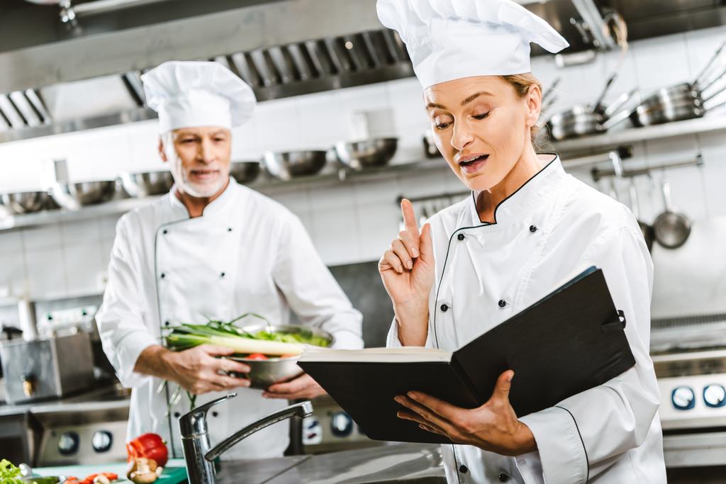 θηλυκό σεφ στην ομοιόμορφη κρατώντας το βιβλίο συνταγών και να κάνει την χειρονομία ιδέα ενώ συνάδελφος μαγείρεμα σε φόντο σε κουζίνα εστιατορίου - Φωτογραφία, εικόνα