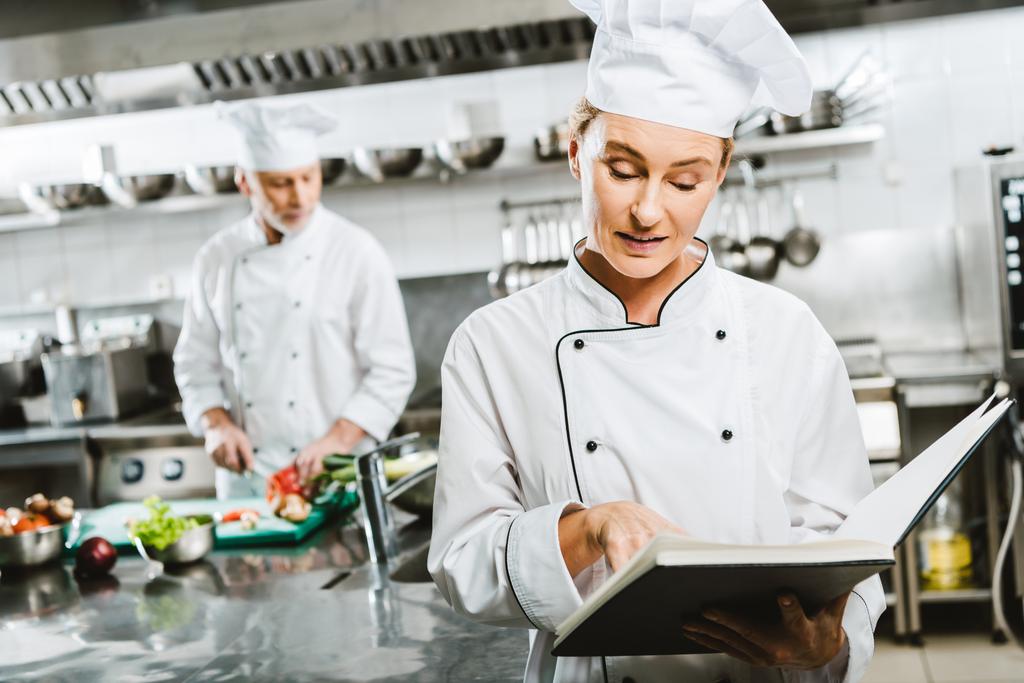 жіночий шеф-кухар в уніформі читання книга рецептів, в той час як колега готує на фоні кухні ресторану
 - Фото, зображення
