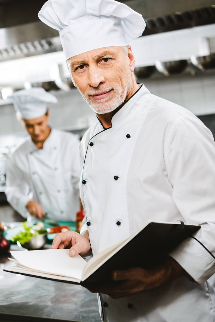 Koch in Uniform und Hut blickt in die Kamera und hält in der Restaurantküche Rezeptbuch in der Hand - Foto, Bild