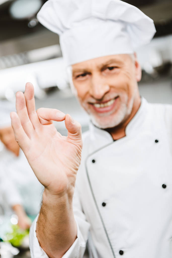 guapo sonriente macho chef en uniforme mirando cámara y mostrando ok signo en restaurante cocina
 - Foto, imagen